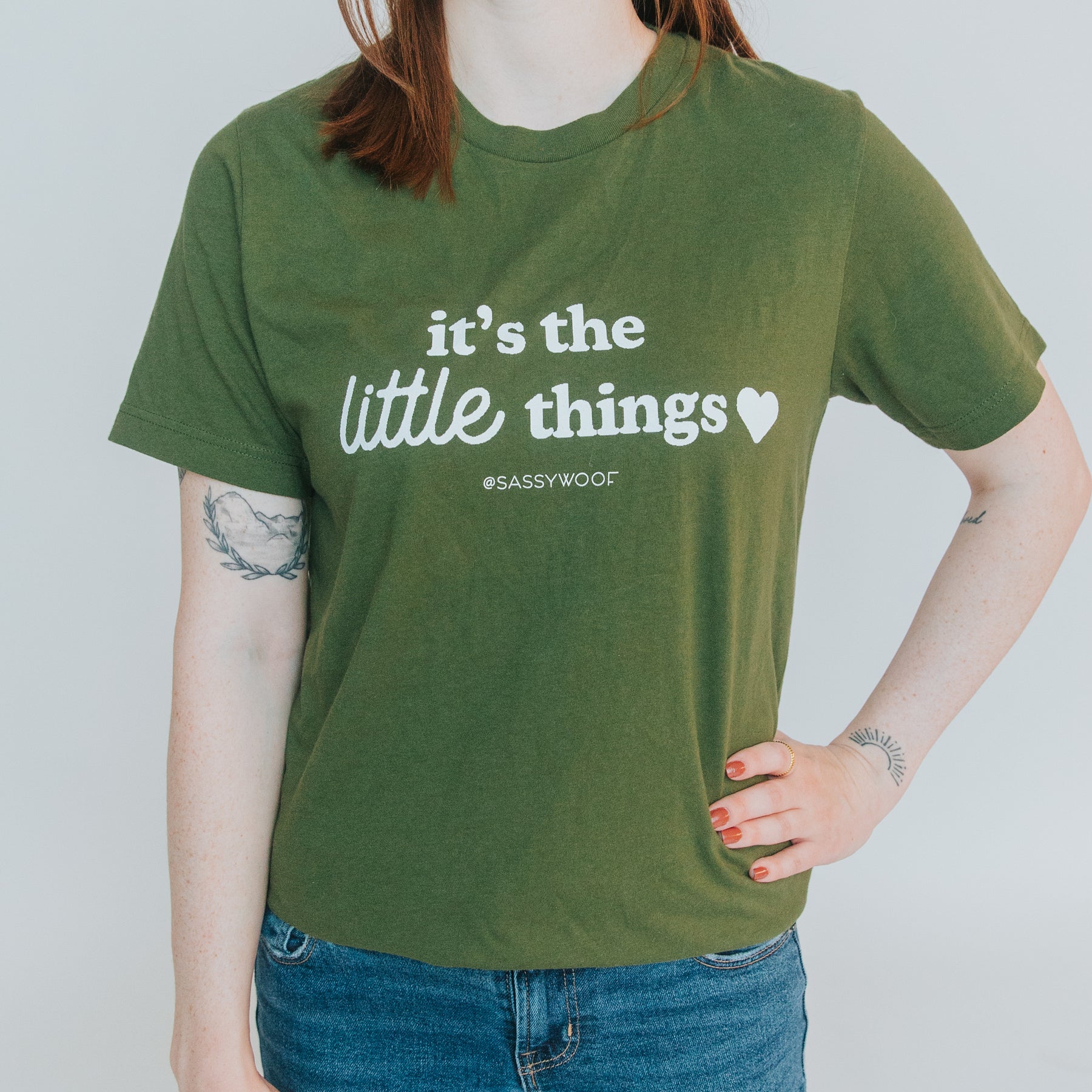 A red-haired person with tattoos stands against a plain background wearing the Human Tee - It’s the Little Things by Sassy Woof, a green, lightweight unisex 100% ring-spun cotton t-shirt, paired with blue jeans.