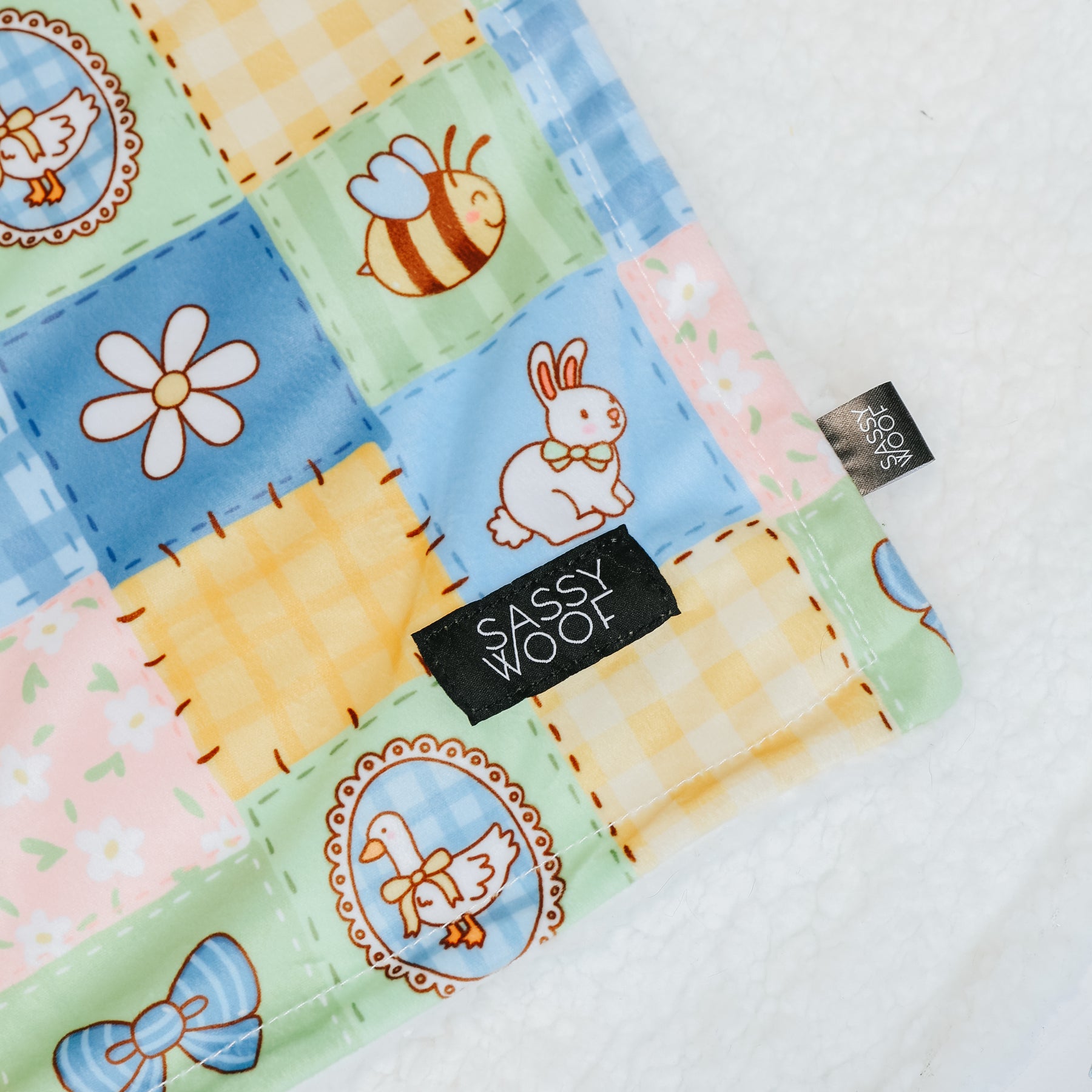 A close-up of the Patch Perfect Pet Blanket by Sassy Woof, showcasing a picnic-themed quilted pattern with bees, bunnies, daisies, ducks, and bows. Made from soft polyester with a black "Sassy Woof" label, displayed on a plush white surface.