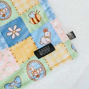 A close-up of the Patch Perfect Pet Blanket by Sassy Woof, showcasing a picnic-themed quilted pattern with bees, bunnies, daisies, ducks, and bows. Made from soft polyester with a black "Sassy Woof" label, displayed on a plush white surface.