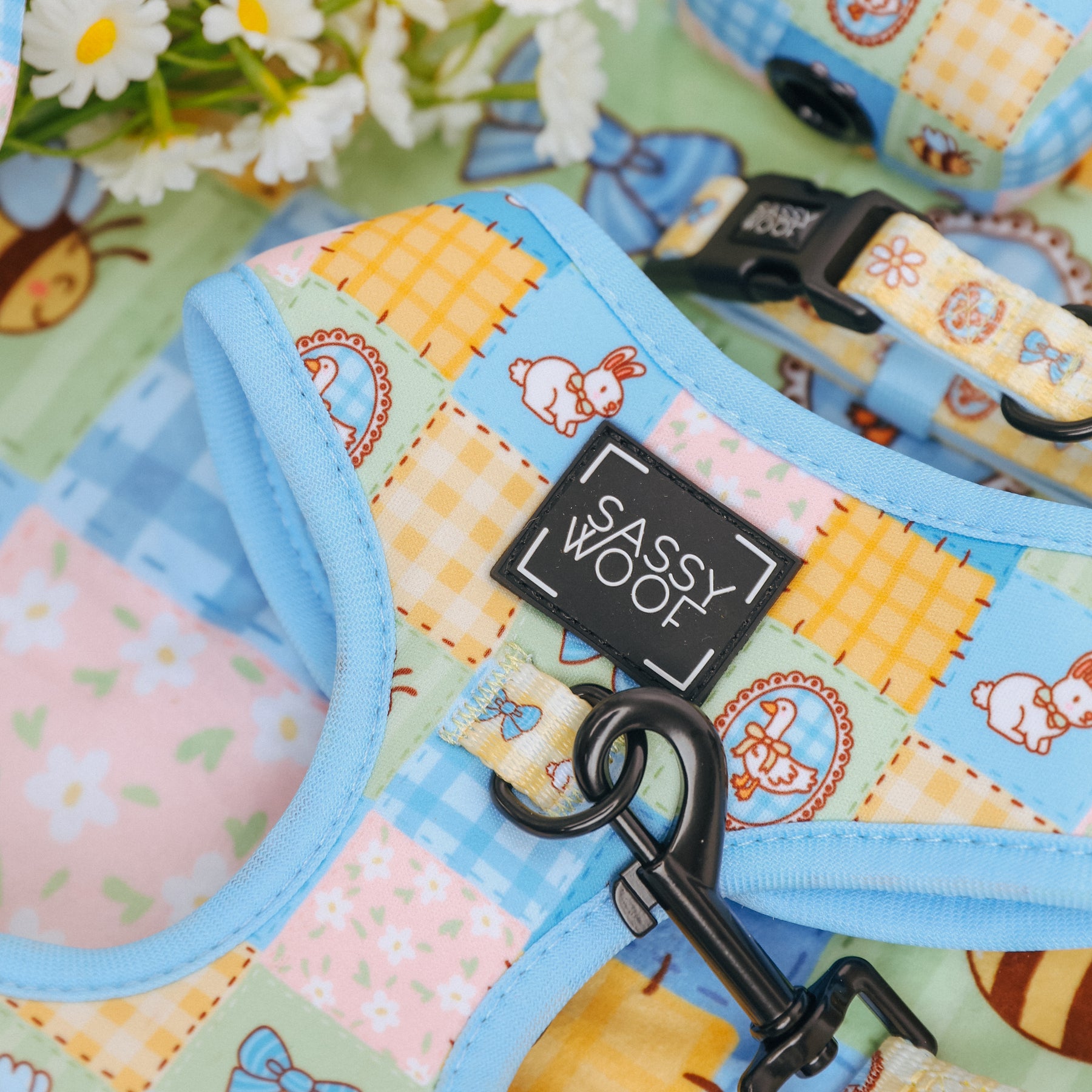 A close-up of the Sassy Woof Dog Adjustable Harness - Patch Perfect, featuring a vibrant patchwork with bunnies and flowers, blue trim, and a branded label. White daisies peek out behind it. Ideal for your puppy’s growth plan.