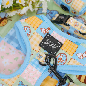 A close-up of the Sassy Woof Dog Adjustable Harness - Patch Perfect, featuring a vibrant patchwork with bunnies and flowers, blue trim, and a branded label. White daisies peek out behind it. Ideal for your puppy’s growth plan.