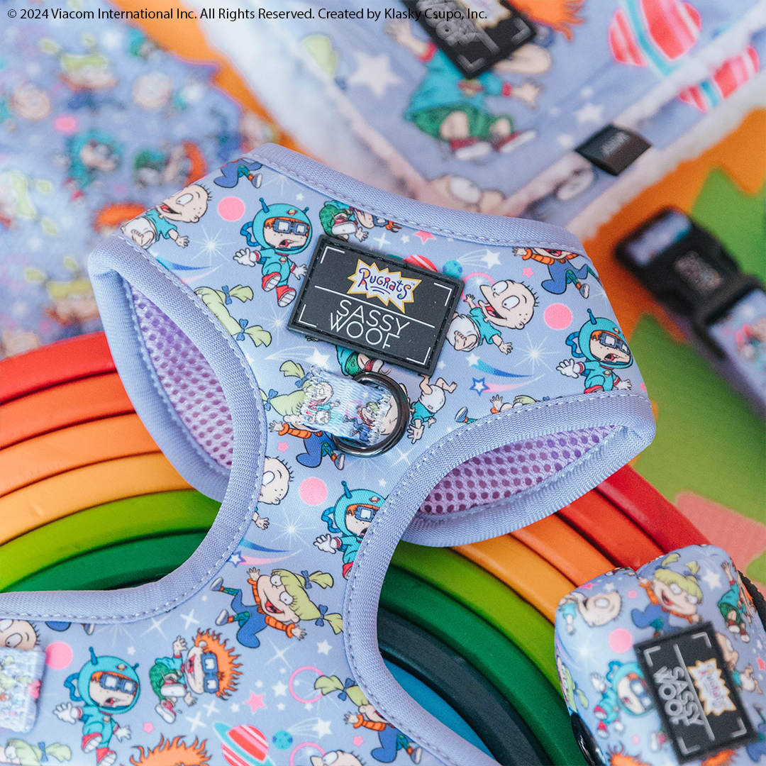 A close-up of the Sassy Woof Dog Adjustable Harness - Nickelodeon Rugrats™ in pastel purple, displayed on a colorful rainbow stack and surrounded by matching dog accessories.