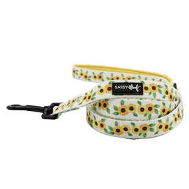 The Sassy Woof "Dog Leash - Sunflower Dreams" features green leaves and yellow sunflowers on white, a padded handle, sturdy black metal clip, and the Sassy Woof label—perfect for stylish outdoor adventures with your pup.