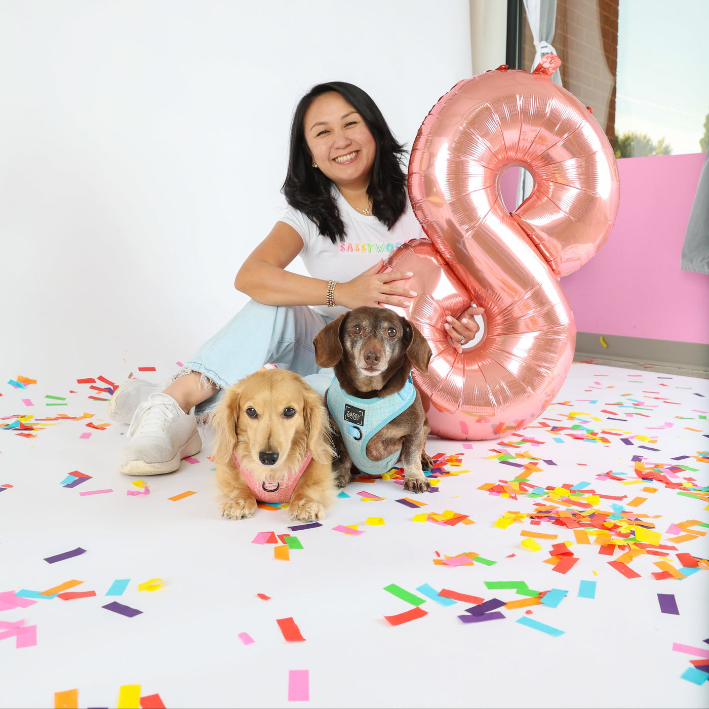 A woman sitting on a white floor with colorful confetti, next to a large metallic pink number 8 balloon, with two small dogs in front of her. The woman is wearing a white t-shirt and light blue jeans, and the dogs are wearing pet accessories.