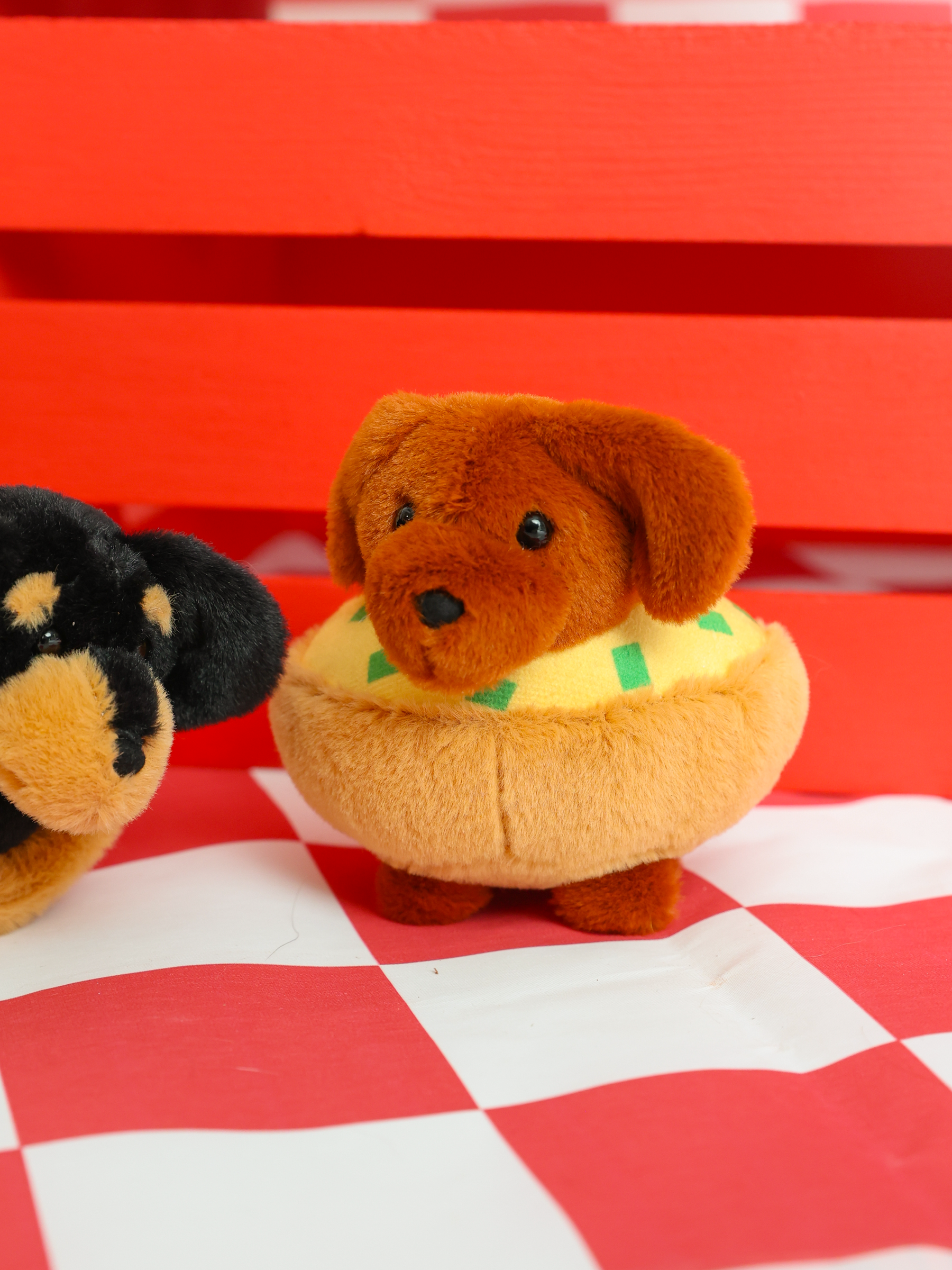 A Sassy Woof Blind Box features a collectible stuffed dog in a yellow bun costume with green relish, posed on a red-and-white checkered surface next to another black and brown plush toy.