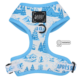 The Sassy Woof Dog Adjustable Harness - Après Ski Club is a light blue and white neoprene harness decorated with winter-themed illustrations, including skiers, mountains, and chalets. It features a black Sassy Woof label, a metal ring for leash attachment, and “Après Ski” printed on the adjustable strap.