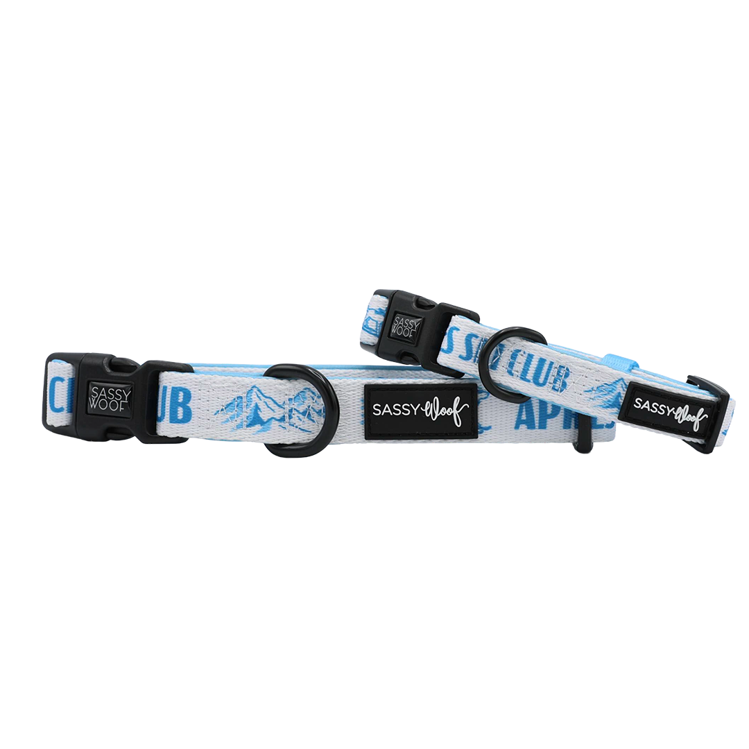 Two adjustable Sassy Woof Après Ski Club dog collars in blue and white with mountain designs, featuring black plastic buckles and D-rings, are displayed against a white background.