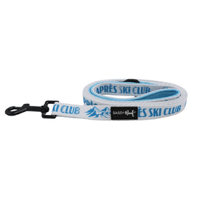 The Sassy Woof Dog Leash - Après Ski Club showcases a white and blue cozy cabin design with bold APRÈS SKI CLUB lettering and a mountain graphic. It features a black metal clasp, a black Sassy Woof label, and a padded neoprene handle for added comfort.