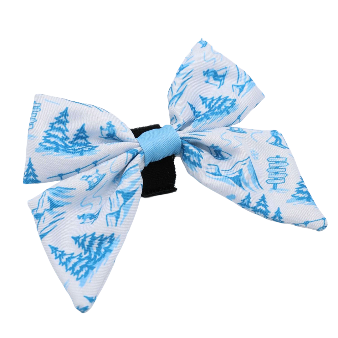 The Dog Sailor Bow - Après Ski Club by Sassy Woof features a white bow with a blue elastic center, adorned with winter-themed blue illustrations of trees, mountains, and animals. Attached to a black clasp, it’s the perfect way to add Apres Ski Club style to your dog’s collar or winter accessories collection.