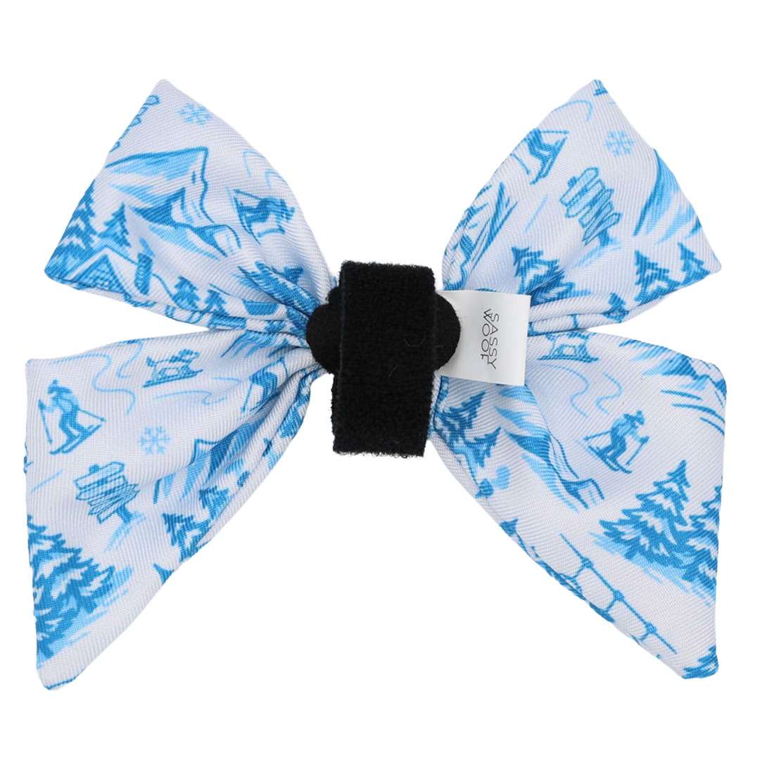 Introducing the Dog Sailor Bow - Après Ski Club by Sassy Woof—a cozy accessory adorned with blue winter-themed prints featuring skiers, snowflakes, trees, and cabins. This charming bow includes a black center band and a white tag, making it the perfect addition to your pup’s collar for all Apres Ski Club adventures.