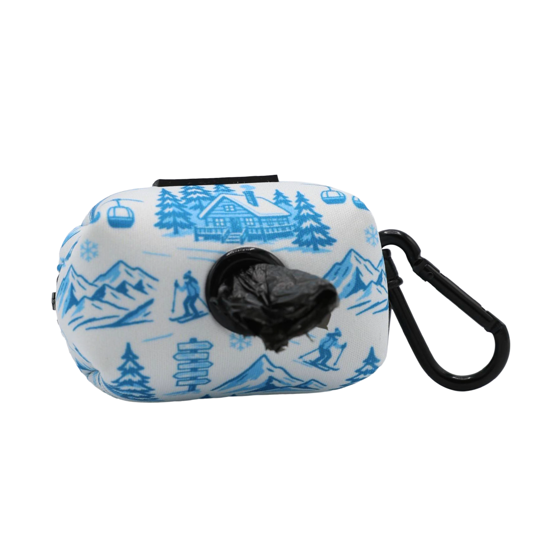Introducing the Dog Waste Bag Holder - Après Ski Club by Sassy Woof: a compact neoprene pouch adorned with blue winter-inspired designs, featuring a front dispenser for black waste bags and a black carabiner clip on the right. This stylish accessory is perfect for adding après ski flair to your dog’s look during daily walks.