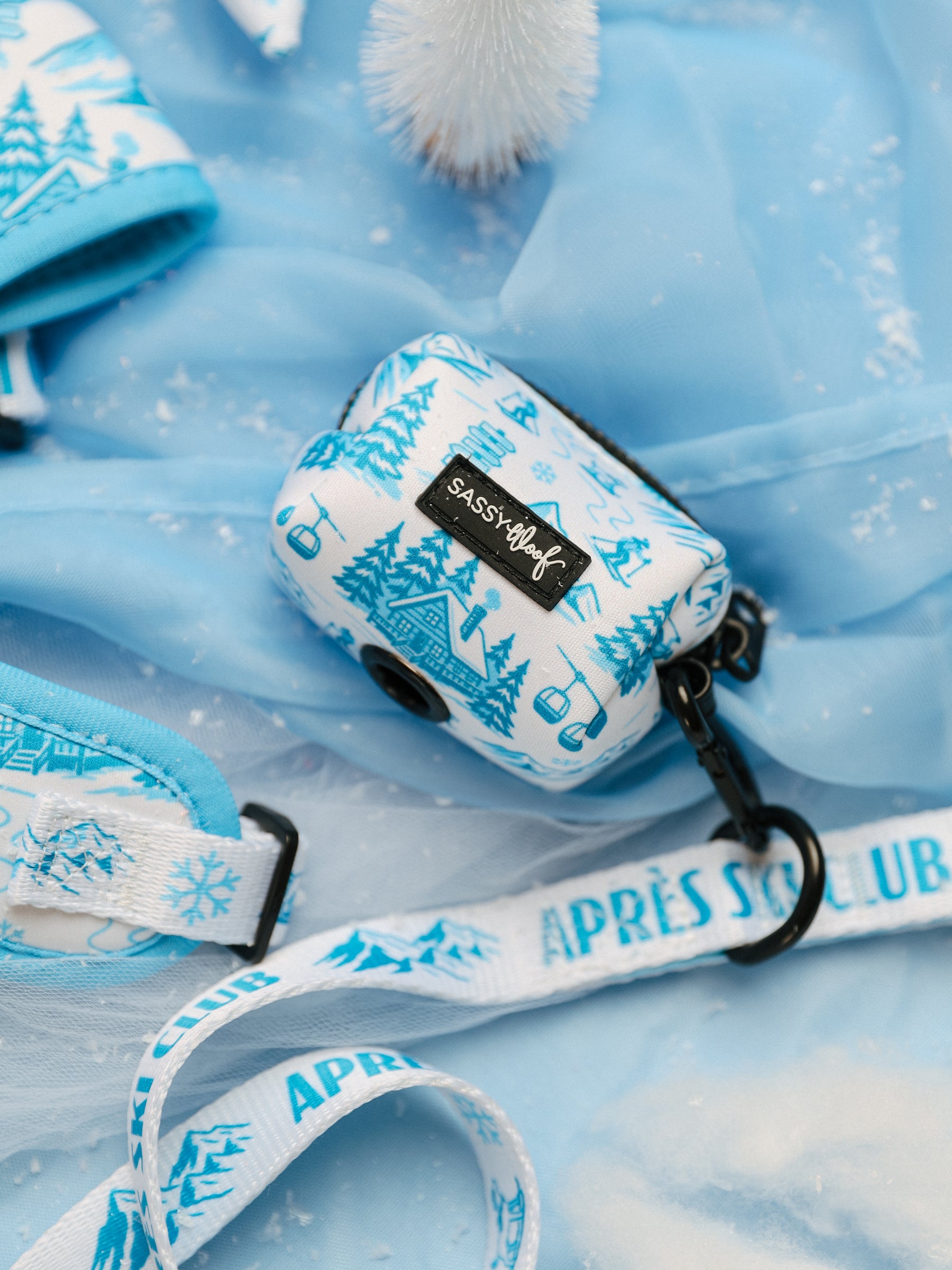 A blue and white patterned neoprene Dog Waste Bag Holder - Après Ski Club from Sassy Woof, featuring ski and tree designs, rests on light blue fabric beside a matching leash labeled “APRÈS SKI CLUB.” This Sassy Woof dog accessory is the perfect way to showcase winter dog fashion, while a small white tree decoration in the background adds a festive touch.
