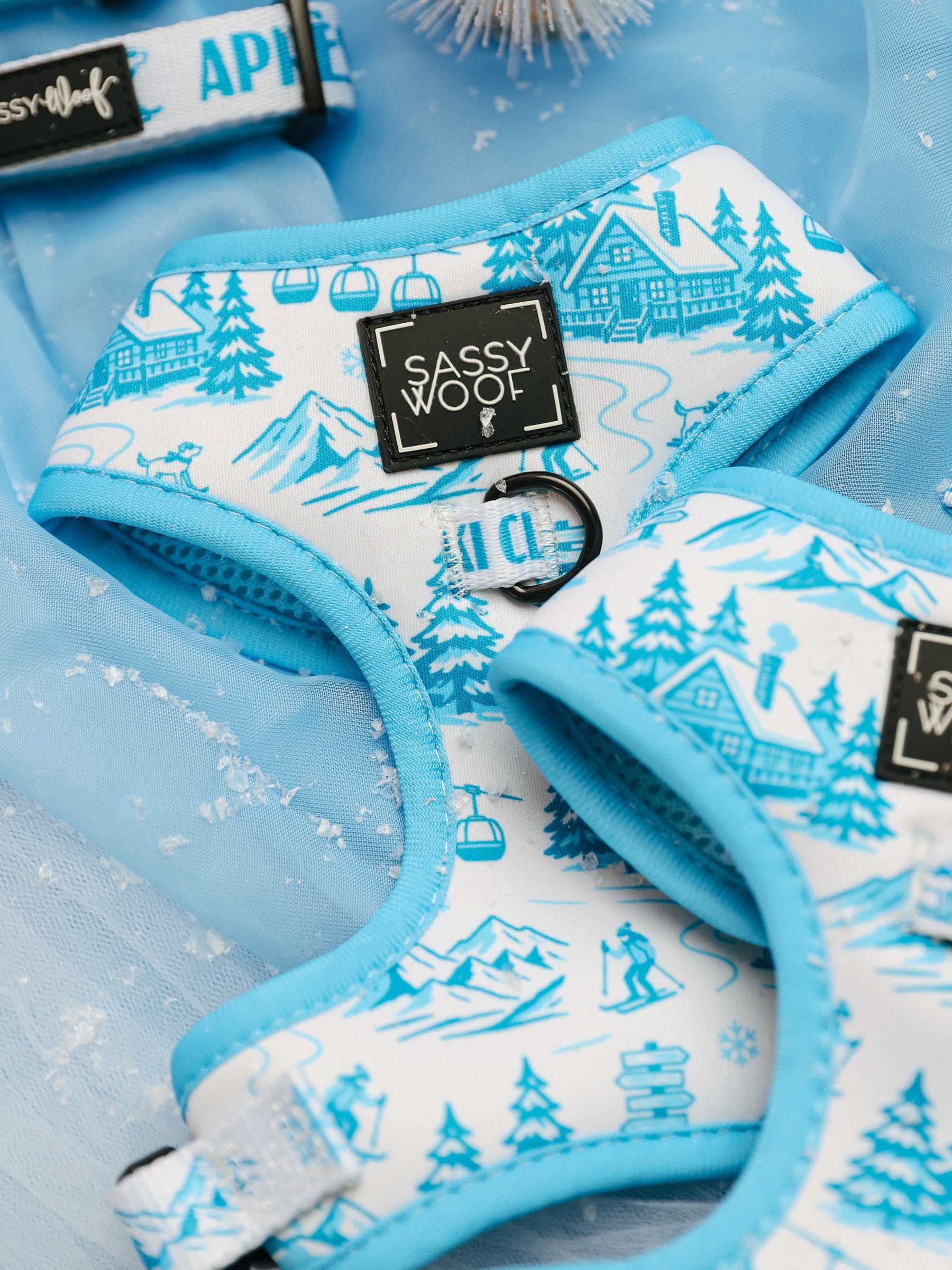Close-up of the Sassy Woof Dog Adjustable Harness - Après Ski Club featuring blue and white custom printed neoprene with skiing and chalet patterns, displayed on light blue fabric with faux snow and part of the APRES leash in view. These adjustable harnesses blend comfort and style for your pup’s winter adventures.