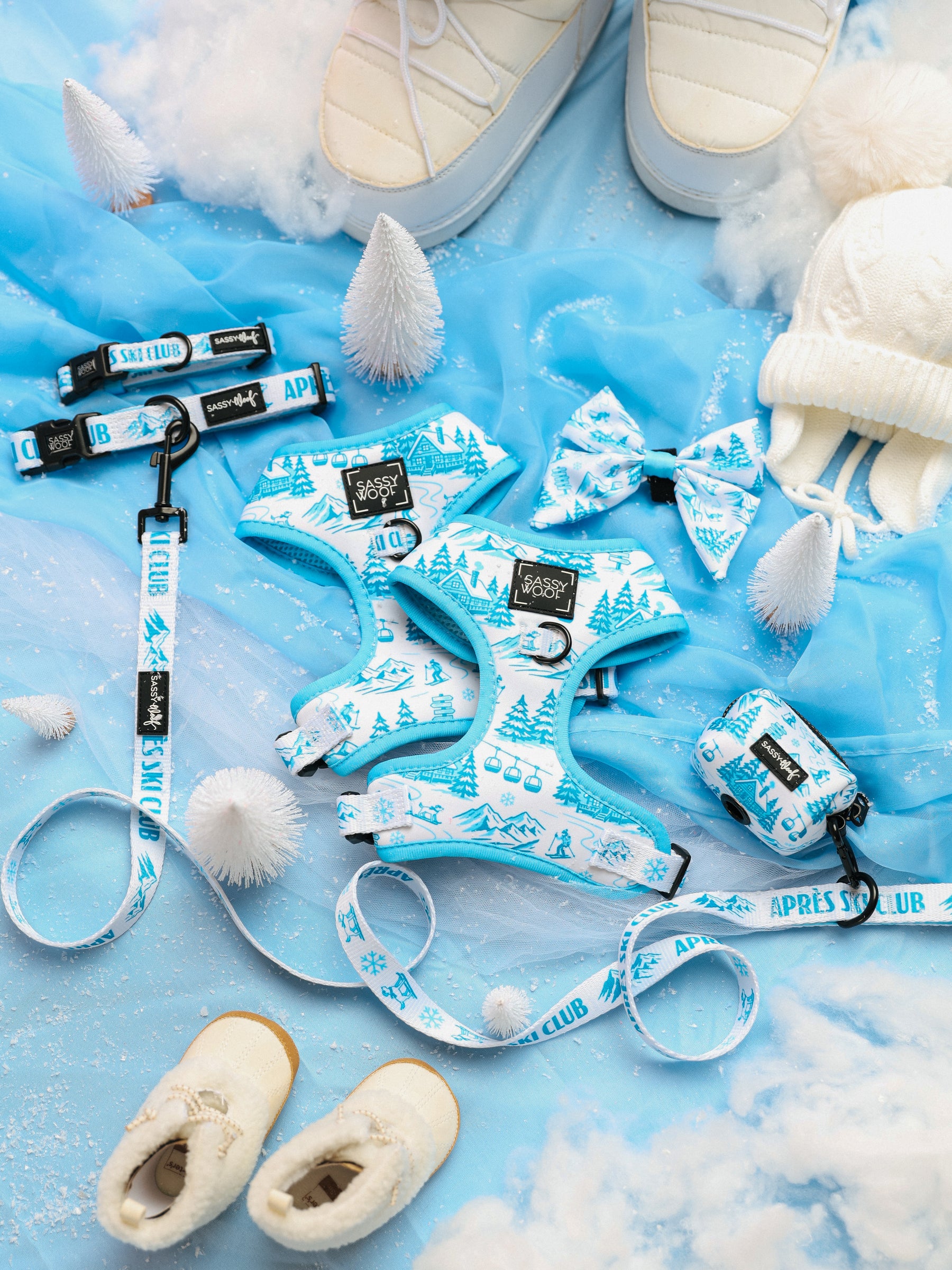 Flat lay of the Sassy Woof Dog Four Piece Bundle - Après Ski Club, featuring adjustable dog collars, dog harnesses, leashes, a bow tie, and a poop bag holder with winter designs, styled alongside white shoes, a knit hat, faux snow, and small white trees on a blue background.