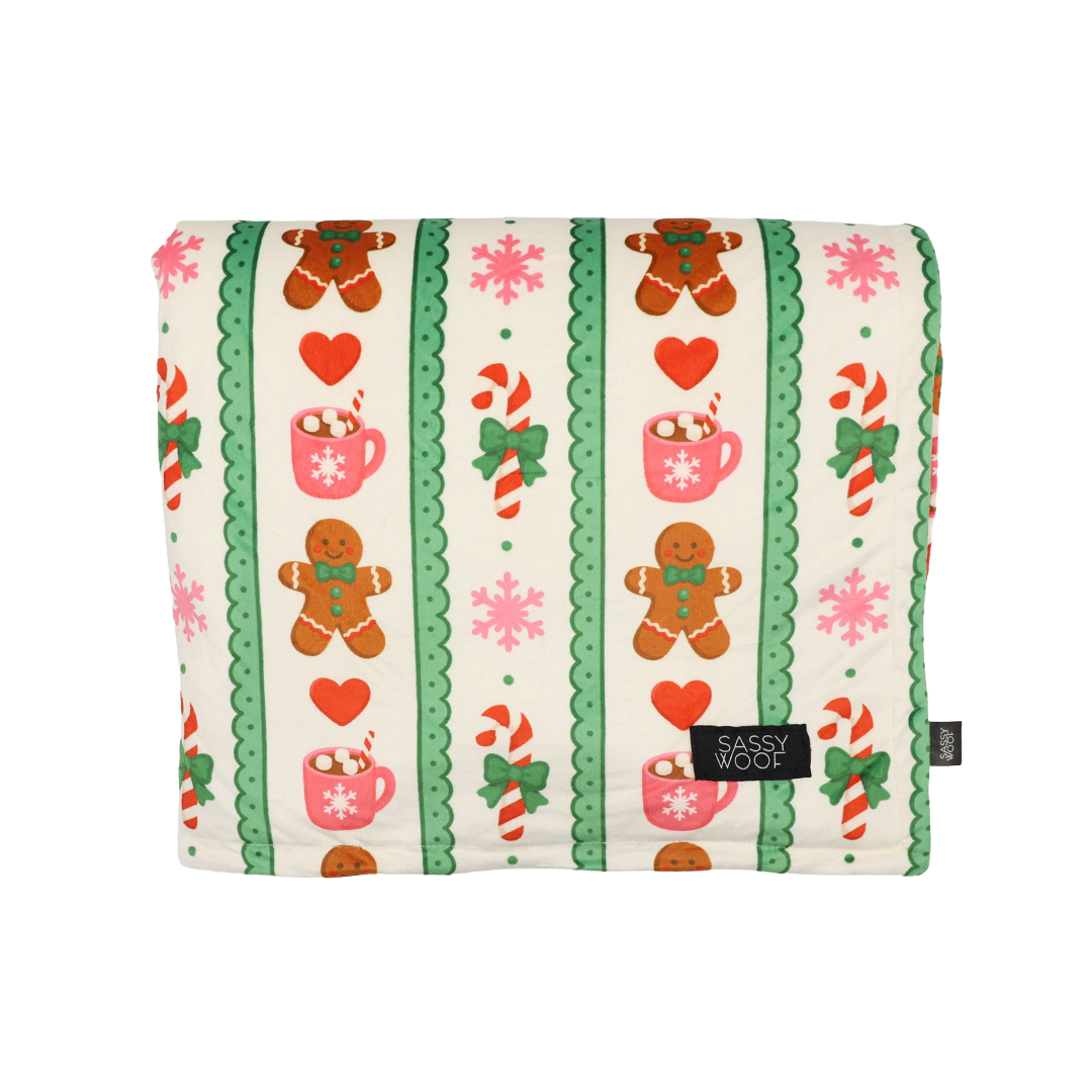 Wrap your furry friend in festive comfort with the Sassy Woof Pet Blanket - Cocoa & Cookies. This cozy winter blanket, part of the Cocoa & Cookies collection, features a cheerful pattern of gingerbread men, hearts, candy canes, pink snowflakes, and holiday mugs set against green and cream stripes. Finished with a black Sassy Woof tag sewn on the front, it makes a thoughtful gift for the holiday season.