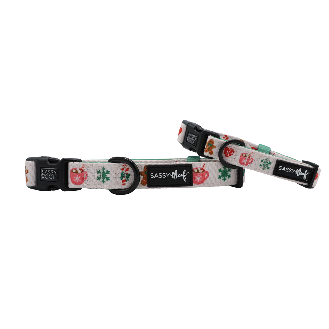 Two Sassy Woof “Cocoa & Cookies” adjustable dog collars featuring festive holiday designs—gingerbread men, snowflakes, and mugs—on a white background. Each neoprene collar includes black plastic buckles and a signature Sassy Woof logo tag.