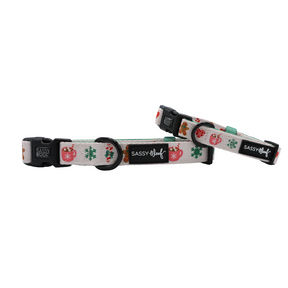 Two Sassy Woof “Cocoa & Cookies” adjustable dog collars featuring festive holiday designs—gingerbread men, snowflakes, and mugs—on a white background. Each neoprene collar includes black plastic buckles and a signature Sassy Woof logo tag.