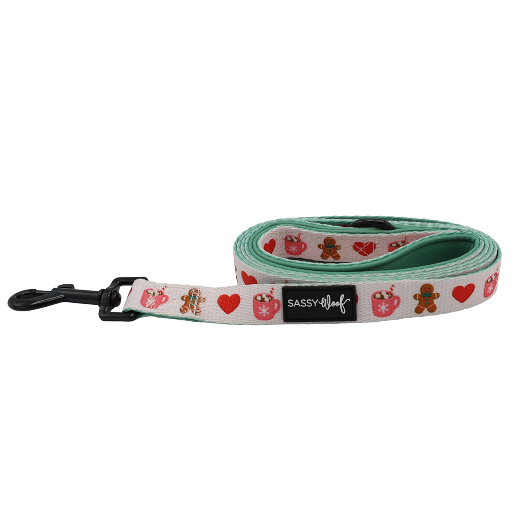 The Dog Leash - Cocoa & Cookies from Sassy Woof features playful images of hearts, gingerbread men, and mugs along its rolled-up design with pink edges. A black clasp and Sassy Woof label add a stylish flair, while the neoprene handle ensures a comfortable grip for every walk.