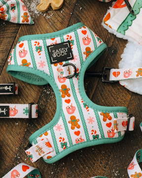 The Sassy Woof Dog Two Piece Bundle - Cocoa & Cookies features an adjustable dog harness with green trim and festive holiday patterns such as gingerbread men, hearts, and candy canes. Shown on a wooden surface alongside cheerful holiday dog accessories, this harness is part of the charming Cocoa & Cookies collection.