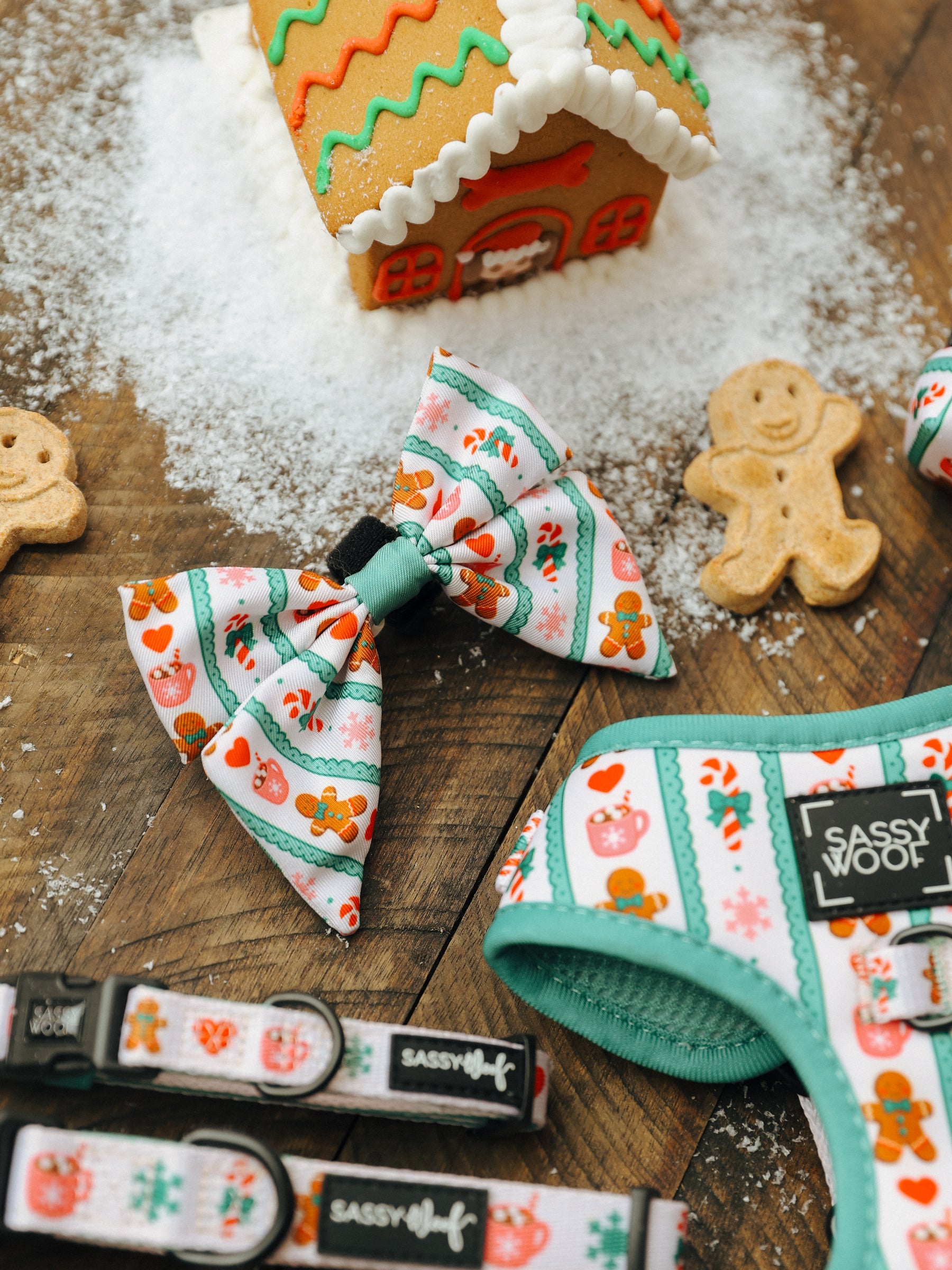 Sassy Woof’s Dog Sailor Bow from the Cocoa & Cookies collection, adorned with gingerbread and holiday designs, is displayed on a wooden surface beside a gingerbread house, two gingerbread cookies, and a dusting of powdered sugar. This festive accessory adds holiday cheer to your pet’s look for the season.