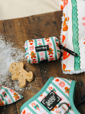 A Sassy Woof Dog Waste Bag Holder from the Cocoa & Cookies collection, featuring gingerbread and candy designs, sits on a wooden table beside a gingerbread cookie, sprinkled flour, and matching patterned items like an adjustable harness.