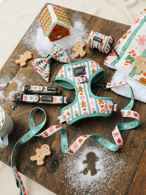 Sassy Woof’s Dog Four Piece Bundle from the Cocoa & Cookies collection is arranged on a wooden board with gingerbread cookies, faux snow, and a small gingerbread house. The bundle features festive holiday accessories—including an adjustable harness, leash, collar, and bowtie—with charming gingerbread designs.