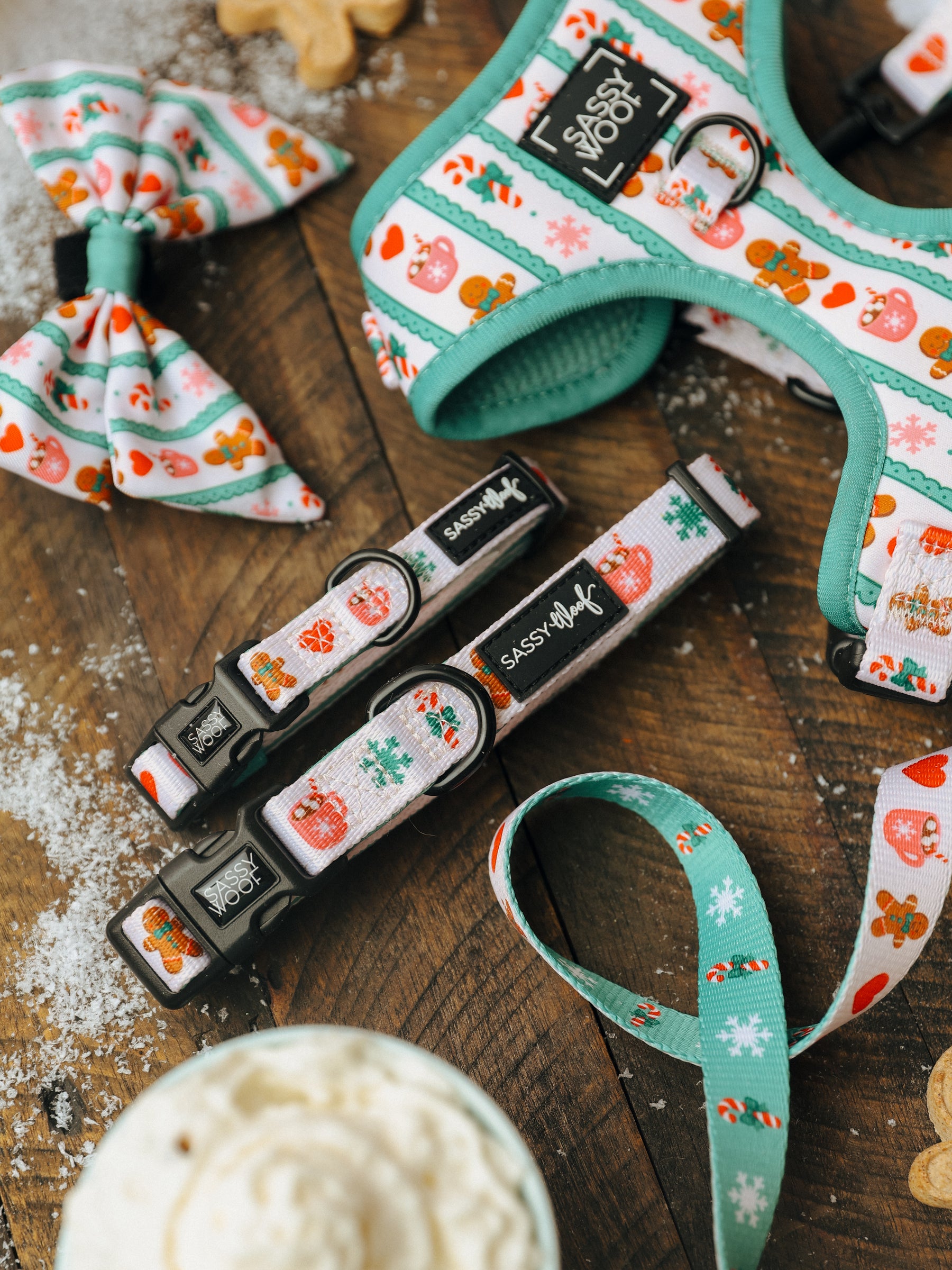 Sassy Woof’s “Dog Collar - Cocoa & Cookies” and matching festive accessories featuring gingerbread and heart motifs—including a bow, harness, two adjustable collars, and a leash—are displayed on a wooden surface dusted with snow-like powder, next to a cozy whipped cream drink.