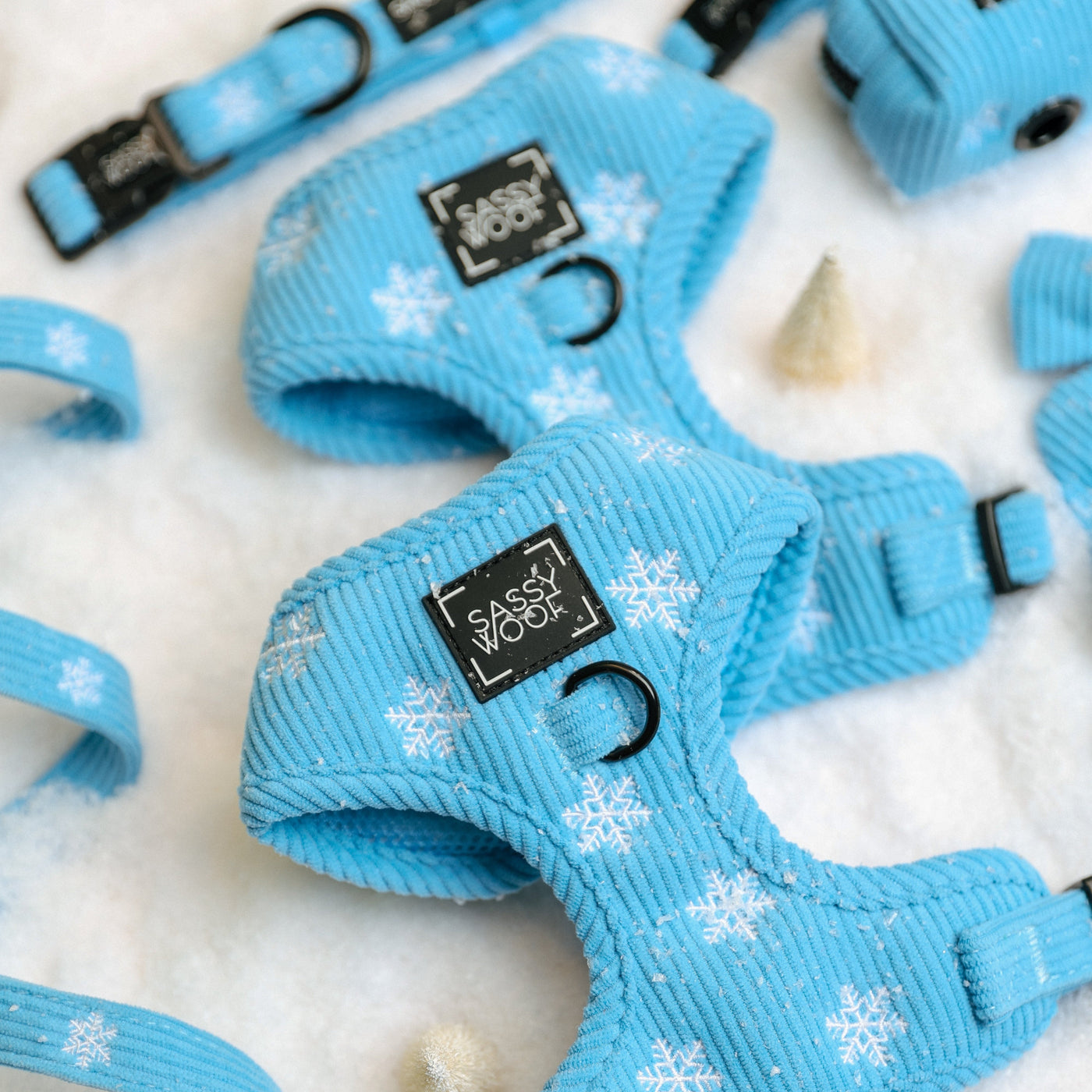 Sassy Woof Dog Four Piece Bundle - Morning Frost featuring a light blue adjustable harness, collar, leash, and waste bag holder with a white snowflake pattern, arranged on a white snowy surface.