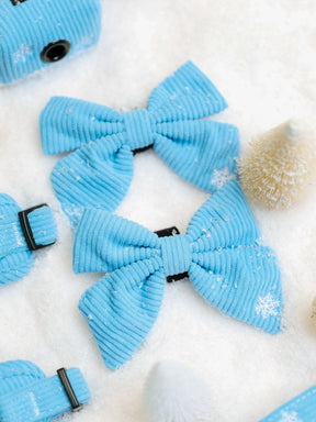 Two light blue corduroy Dog Sailor Bow - Morning Frost bows featuring white snowflake patterns on a snowy surface.