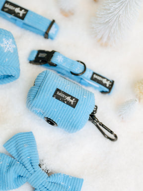 A light blue Sassy Woof Dog Collar Three Piece Bundle - Morning Frost featuring a corduroy snowflake pattern, a waste bag dispenser, and a bow tie, arranged on a snowy surface.