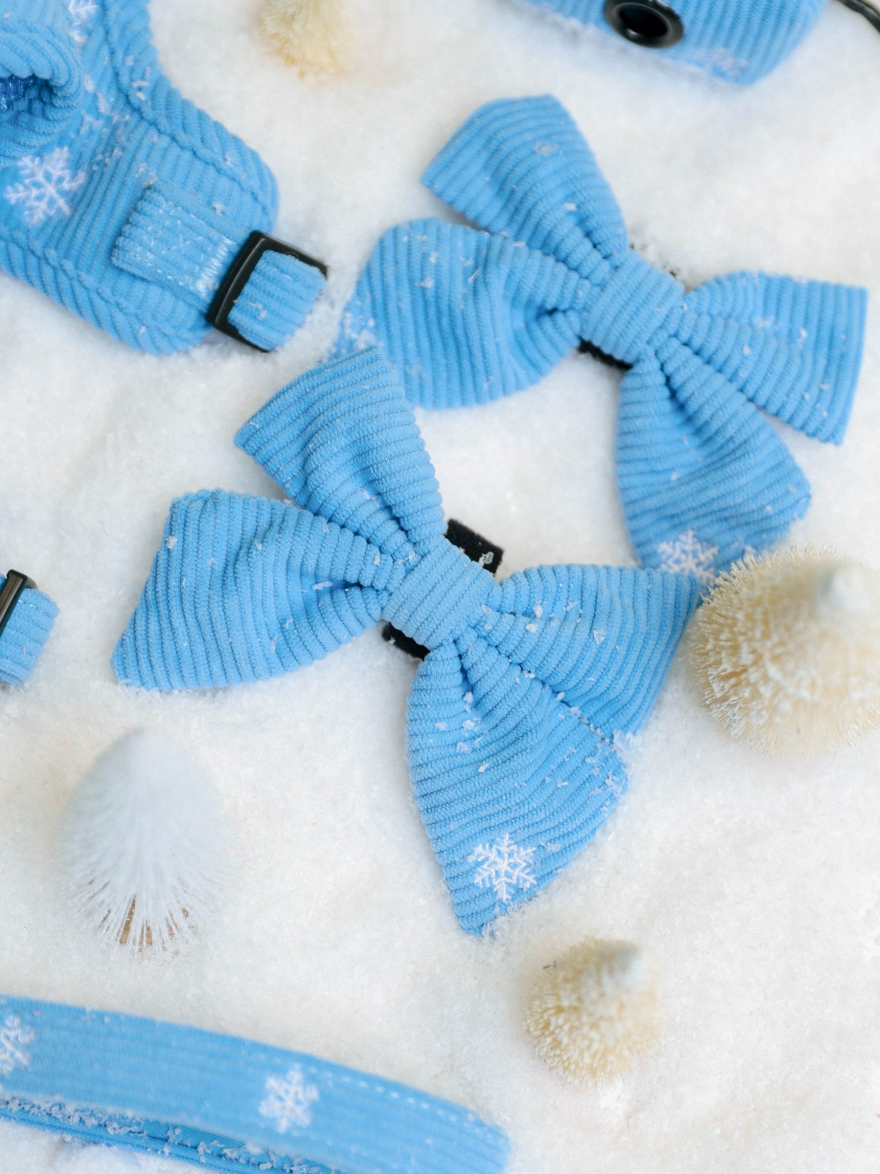 Sassy Woof Dog Sailor Bow - Morning Frost featuring light blue corduroy with white snowflake patterns, displayed on a snowy surface alongside miniature pine trees.