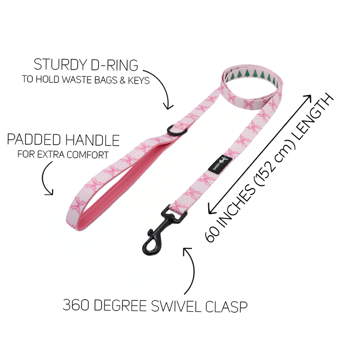 The Sassy Woof Dog Four Piece Bundle - Pretty in Pine includes a pink dog leash featuring a padded handle, sturdy D-ring, 60-inch (152 cm) length, and a 360-degree swivel clasp. The leash is adorned with a charming white pattern of pink bows and small green trees—ideal for complementing your favorite winter dog accessories or pairing with the Pretty in Pine dog harness. Labels clearly highlight each feature.