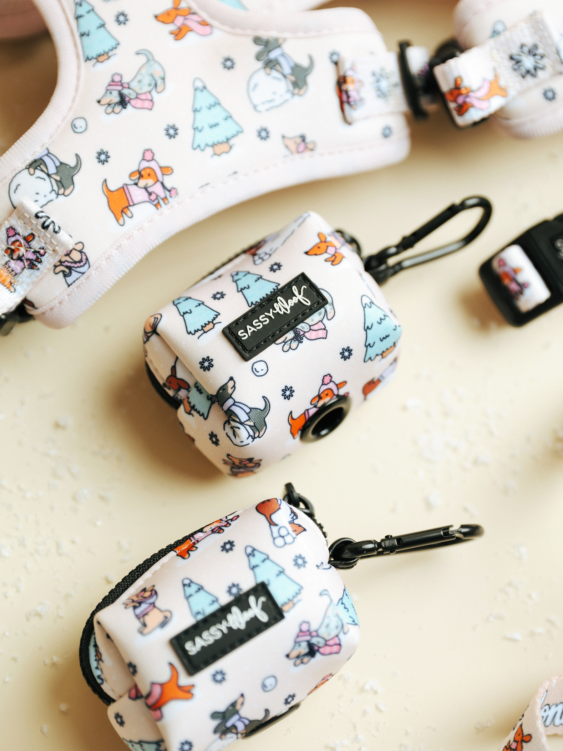 The Weenie Wonderland Collection by Sassy Woof features playful patterned dog accessories on a pale background, including the Dog Waste Bag Holder – Weenie Wonderland with convenient clips. Showcasing lively dog, fox, and tree illustrations, these pieces make perfect winter accessories for dachshunds.