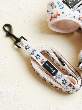 A pastel-colored Dog Leash - Weenie Wonderland from Sassy Woof, featuring playful cartoon dog and tree prints, is coiled on a light surface beside a matching harness. Snow-like flakes are scattered nearby, making these pastel accessories ideal for winter walks.