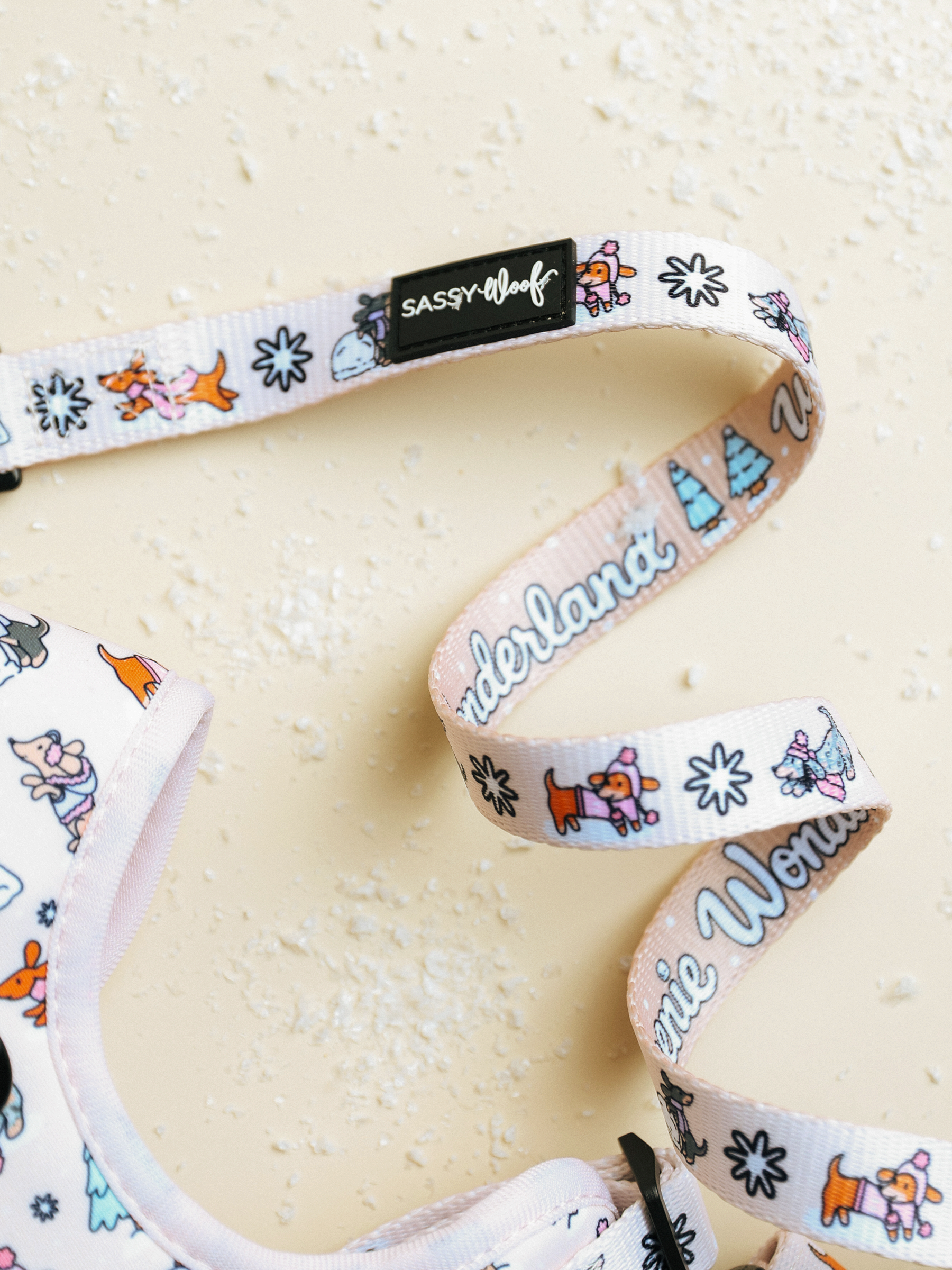 The Dog Leash - Weenie Wonderland from Sassy Woof, featuring playful dog and tree designs along with the word “Wonderland” and a black SASSY WOOF tag, rests on a light beige textured surface dusted with powder. This delightful white leash is ideal for anyone who enjoys pastel-themed dog accessories.