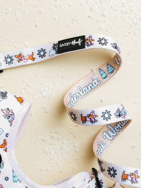 The Dog Leash - Weenie Wonderland from Sassy Woof, featuring playful dog and tree designs along with the word “Wonderland” and a black SASSY WOOF tag, rests on a light beige textured surface dusted with powder. This delightful white leash is ideal for anyone who enjoys pastel-themed dog accessories.