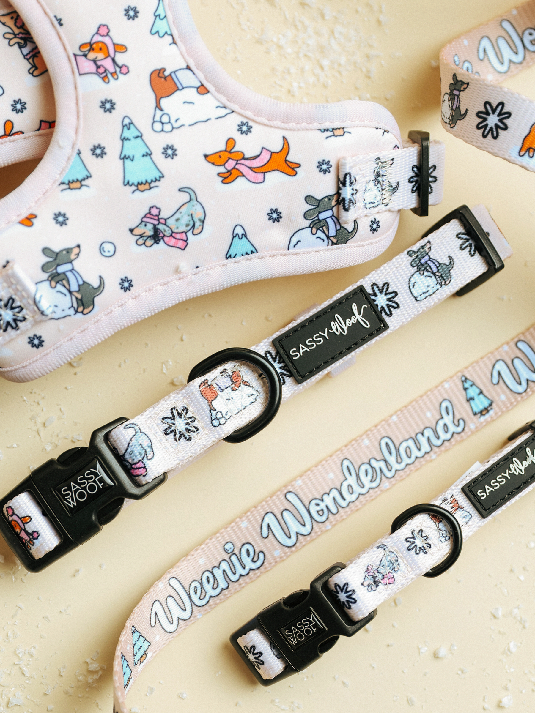 Dog collars from Sassy Woof, including the “Weenie Wonderland” design, feature playful winter-themed cartoon dogs and trees in pastel colors with phrases like “Sassy Woof” and “Weenie Wonderland” on the fabric. Arranged on a light-colored surface, these festive accessories—such as the adjustable Sassy Woof dog collar and the charming Weenie Wonderland collar—are perfect for bringing seasonal cheer to your pup’s wardrobe.