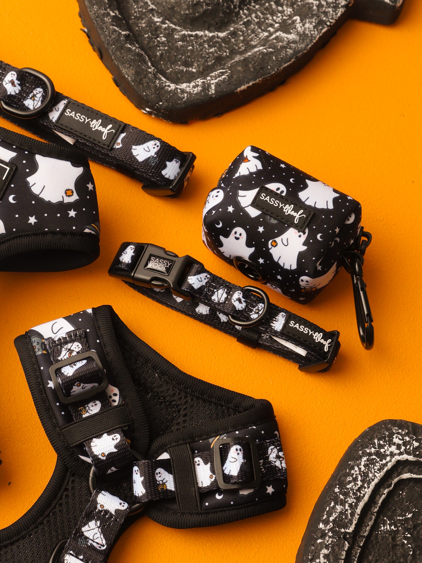 Sassy Woof’s Dog Three Piece Bundle - Lil Boo Thing, featuring black pet accessories with white ghost patterns—a Halloween dog collar, harness, and small pouch—is displayed on an orange surface with black textured objects partially visible around the edges.