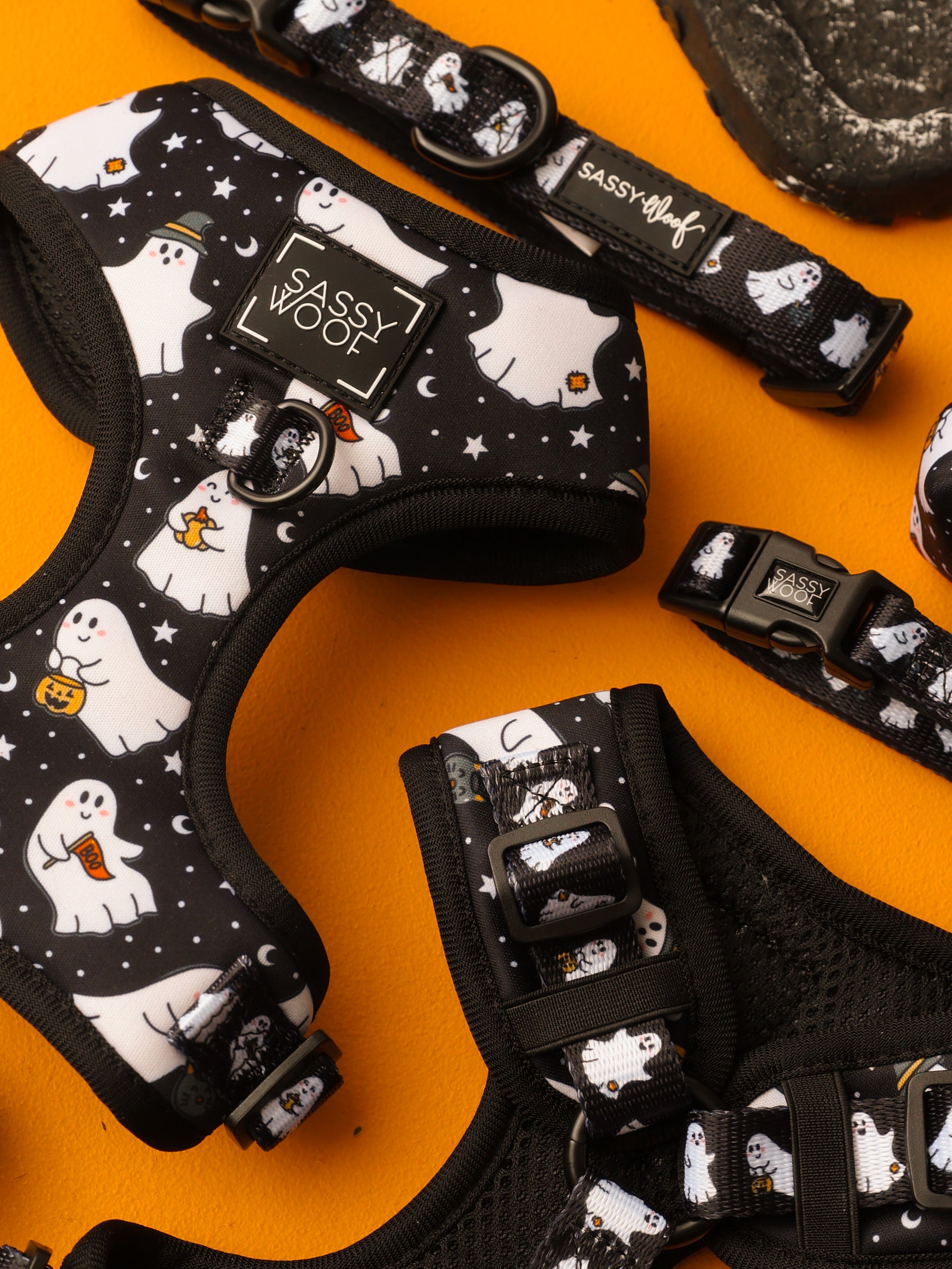 Black dog harnesses and collars from the Sassy Woof Dog Two Piece Bundle - Lil Boo Thing, featuring cute white ghost and star patterns, are displayed on an orange surface. These festive Halloween accessories offer adjustable harness options with playful holiday-themed designs and prominent Sassy Woof labels.