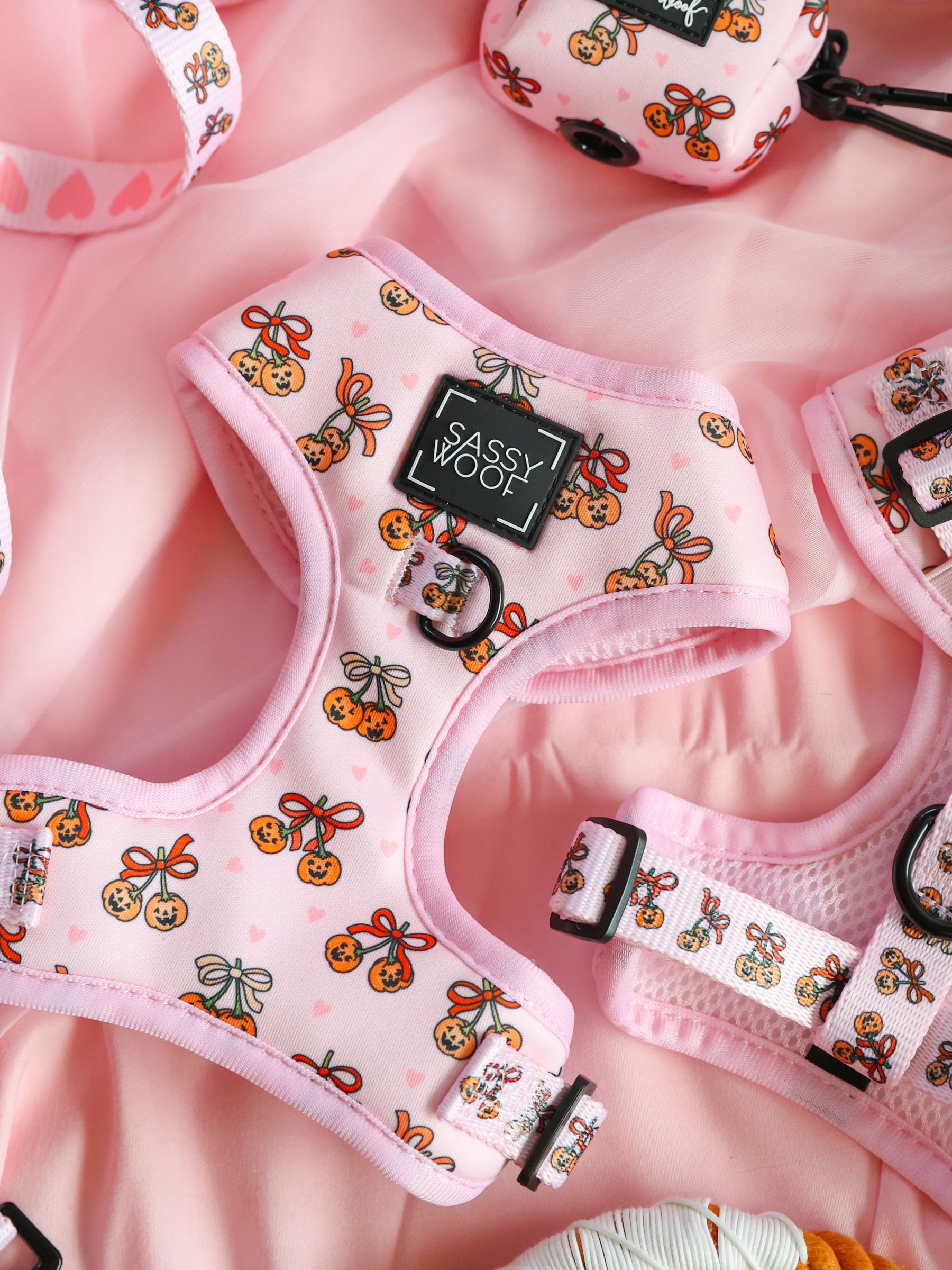 The Sassy Woof Dog Adjustable Harness - Sassy Lil Pumpkins, featuring a cute orange and bow pattern, is displayed on a pink fabric background alongside matching accessories.