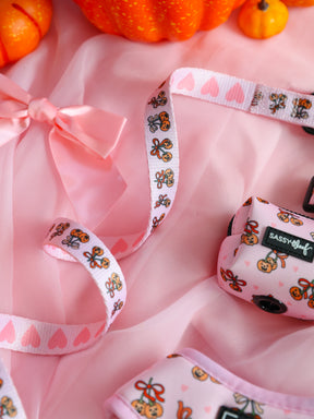 A pink fabric background with a satin bow, orange pumpkins, and the Sassy Woof Dog Leash - Sassy Lil Pumpkins, decorated with gingerbread and heart patterns from the Pumpkins Dog Collection. This leash features a soft neoprene handle for added comfort and pairs perfectly with an adjustable harness for your pup.