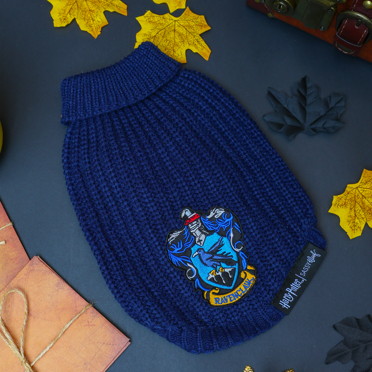 A Sassy Woof Pet Cable Knit Sweater - Harry Potter™ Ravenclaw, featuring a blue cable knit design with the iconic Ravenclaw crest patch, rests on a dark surface surrounded by yellow leaves, a brown book, and a small chest—making it the ideal cozy sweater for your dog during cool autumn days.