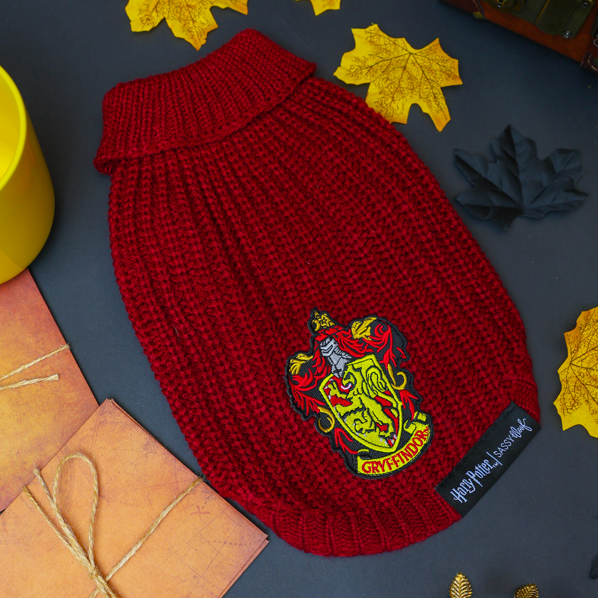 The Sassy Woof Pet Cable Knit Sweater - Harry Potter™ Gryffindor, featuring a vibrant red knit and the iconic Gryffindor crest, is artfully displayed among autumn leaves, a yellow cup, a chest, and wrapped gifts on a dark background—capturing the cozy charm of this magical dog sweater for your furry companion.