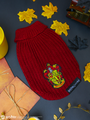 The Sassy Woof Pet Cable Knit Sweater - Harry Potter™ Gryffindor, featuring a vibrant red knit and the iconic Gryffindor crest, is artfully displayed among autumn leaves, a yellow cup, a chest, and wrapped gifts on a dark background—capturing the cozy charm of this magical dog sweater for your furry companion.