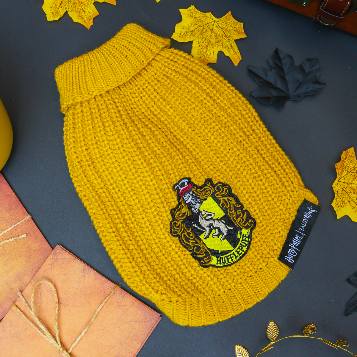 Cuddle your pup in the Pet Cable Knit Sweater - Harry Potter™ Hufflepuff by Sassy Woof, a yellow cable knit dog sweater featuring the iconic Hufflepuff crest patch. Surrounded by autumn leaves, a wooden chest, and wrapped gifts on a dark background, this officially licensed sweater is perfect for any proud Hufflepuff pet.