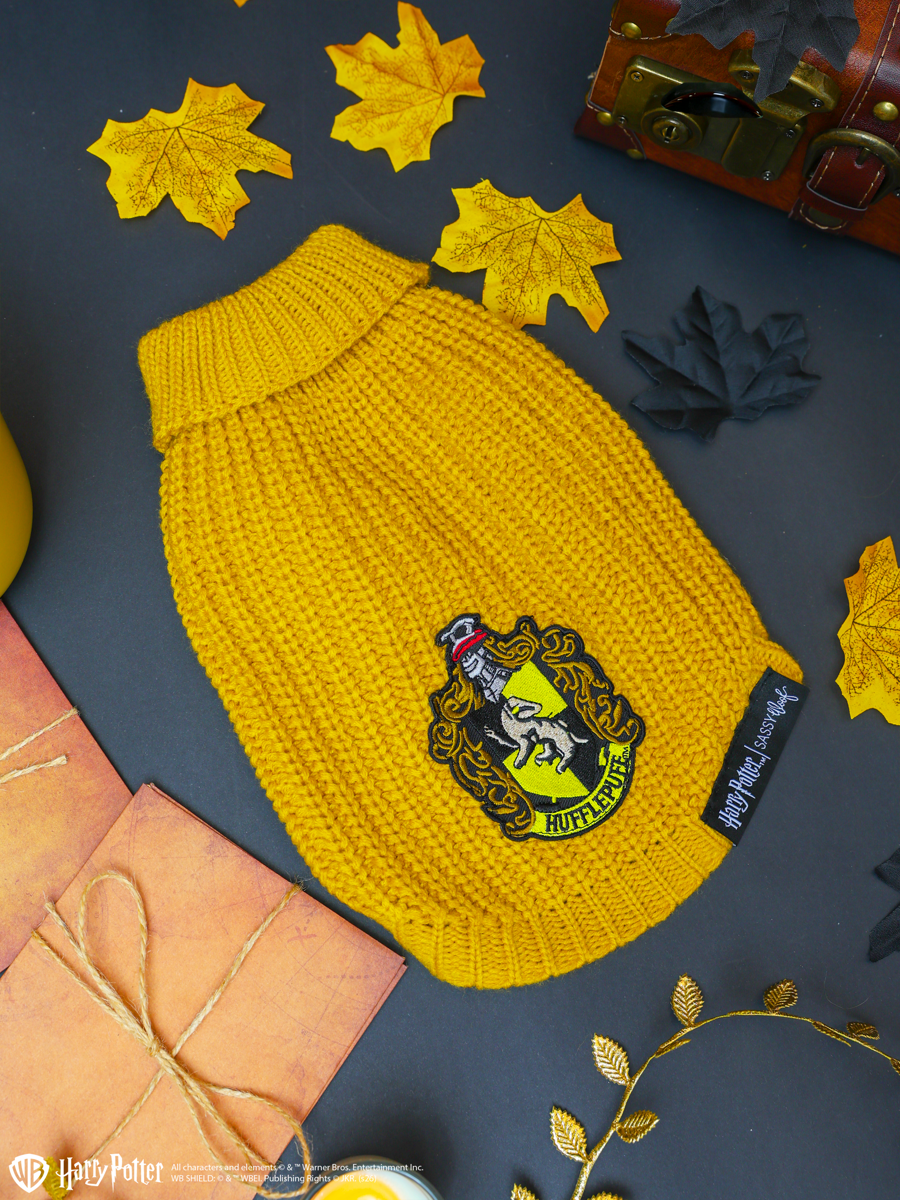 Cuddle your pup in the Pet Cable Knit Sweater - Harry Potter™ Hufflepuff by Sassy Woof, a yellow cable knit dog sweater featuring the iconic Hufflepuff crest patch. Surrounded by autumn leaves, a wooden chest, and wrapped gifts on a dark background, this officially licensed sweater is perfect for any proud Hufflepuff pet.