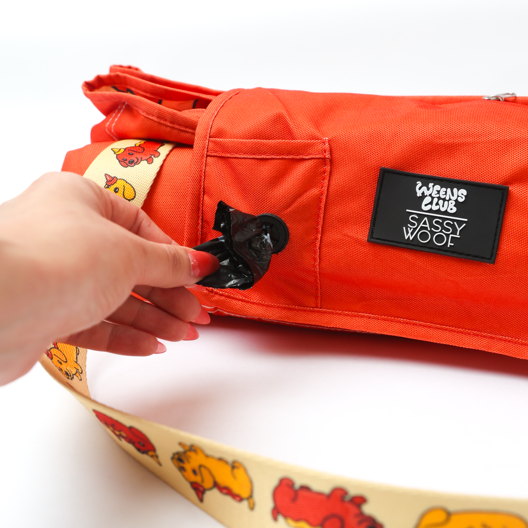 A hand pulls a black waste bag from the dispenser pocket of the orange Sassy Woof Dog Picnic Blanket - Hot Dawg, featuring a cream strap with colorful dachshund designs.