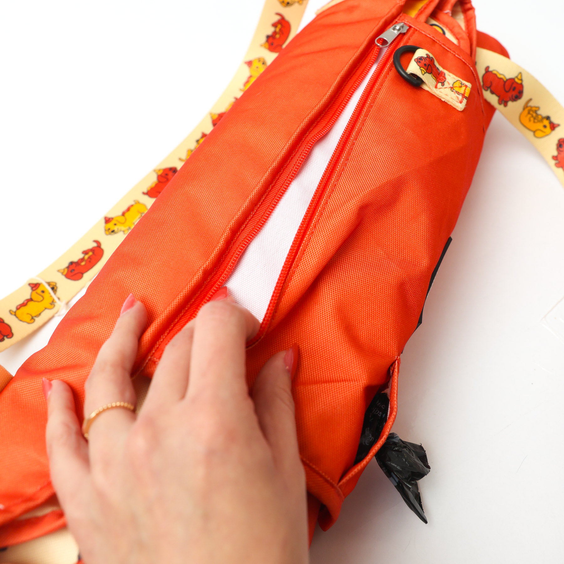 A hand opens the zippered pocket of Sassy Woof’s Dog Picnic Blanket - Hot Dawg, featuring a yellow animal-print strap. Inside, a white lining and black waste bags are visible.