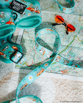 The Sassy Woof Dog Leash – Peanuts™ Road Trippin' features playful Snoopy designs and bright turquoise color, styled for comfort and fun. Set next to orange sunglasses on a map, it suggests cheerful adventures in the sun.