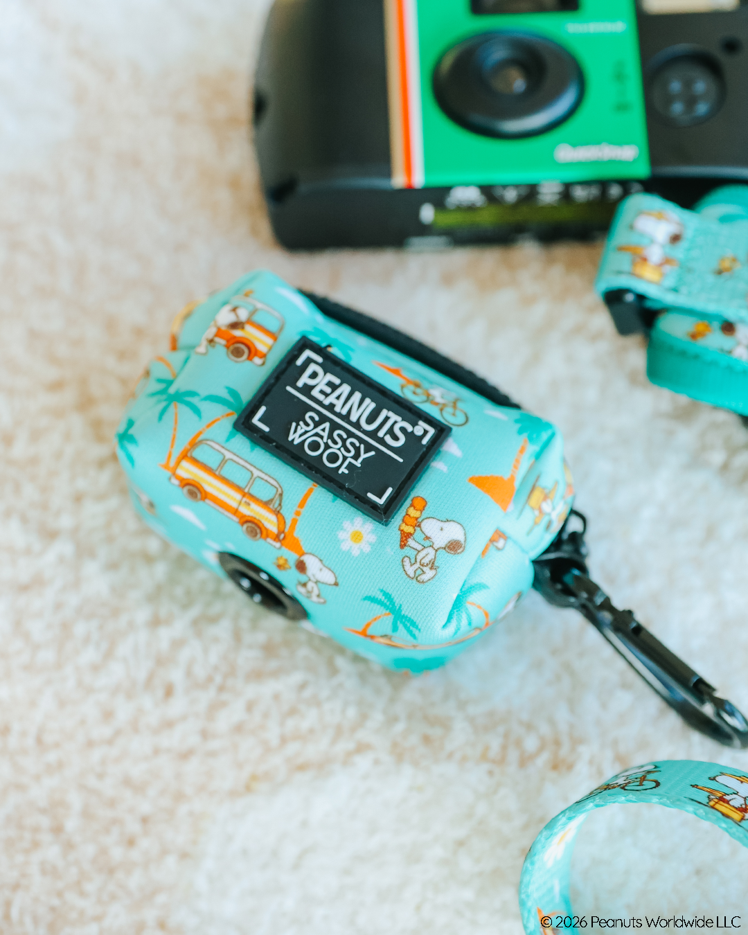 A teal Sassy Woof Dog Waste Bag Holder - Peanuts™ Road Trippin' with Snoopy and van prints rests on a light surface beside a green film camera, matching strap, and adventure-ready dog gear.